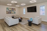 Located at 706 Huffman Mill Rd in Burlington, NC, this single-floor commercial space offers a polished and contemporary environment designed to welcome visitors and support professional activity. The main lounge area serves as the heart of the space, characterized by crisp white walls, warm wood-look flooring, and a drop ceiling outfitted with distinctive diamond-shaped LED light fixtures that provide bright, even illumination throughout. The seating arrangement is thoughtfully composed, featuring neutral-toned sofas and accent chairs arranged around sleek, dark side tables and a textured area rug, creating a comfortable yet refined atmosphere. A wall-mounted flat-screen television anchors one side of the room, flanked by windows with blinds that allow natural light to filter in while maintaining privacy. Decorative framed artwork adorns the walls, adding visual interest and a touch of sophistication. French-style interior doors with glass panes connect the lounge to adjacent areas, including what appears to be a fitness or equipment room visible in the background, suggesting the space supports a range of functional uses. The overall design aesthetic is clean, modern, and professionally appointed, making it well-suited for client-facing or wellness-oriented business operations.