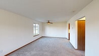 This single-floor home offers a practical and comfortable layout with a clean, neutral aesthetic throughout. The living area is generously sized, featuring soft gray carpeting, warm wood baseboards, and a ceiling fan that anchors the space. Multiple windows allow abundant natural light to fill the room while providing pleasant views of the surrounding greenery and neighborhood.

The galley-style kitchen is well-equipped with warm oak cabinetry lining both sides, light countertops, a stainless steel sink, and white appliances including a range and refrigerator. Tile flooring adds durability and a clean look to the kitchen area, which connects conveniently to other parts of the home through a central hallway.

The bedrooms are carpeted and feature the same neutral wall tones found throughout the home. Warm wood bi-fold closet doors add character and provide ample storage. Each bedroom benefits from windows that bring in natural light and outdoor views.

The bathroom is functional and well-appointed, featuring tile flooring, a vanity with wood cabinetry, and a full tub and shower combination with chrome fixtures and a curtain rod. The consistent use of warm wood tones across cabinetry, doors, and trim gives the home a cohesive and inviting character throughout its single level.