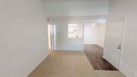 This well-maintained single-floor unit offers a bright, functional layout with a clean, neutral aesthetic throughout. The open living and dining area is finished with plush beige carpeting and features a vaulted ceiling detail, a front entry door, and a large sliding glass door fitted with vertical blinds that invites natural light and outdoor views. The transition between the entry and the living space is marked by wood-look laminate flooring, adding a subtle contrast to the soft carpeted areas.

The galley-style kitchen is efficiently designed with ample white cabinetry lining both walls, light-toned countertops, a double sink, a dishwasher, and a range with a coil cooktop. A skylight-style overhead fixture brightens the space, and wood-look flooring runs throughout, complementing the crisp white finishes. A pass-through window connects the kitchen to the main living area, enhancing the open feel.

The bedroom is a comfortably sized, carpeted retreat with a textured ceiling and a window that frames leafy outdoor greenery, providing a serene atmosphere. The bathroom features a wide vanity with a single sink, a large mirror, a wall-mounted light fixture, built-in storage cabinetry, and a towel bar, all finished in warm neutral tones. The overall ambiance of the unit is clean, bright, and move-in ready.