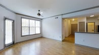 This single-floor space offers a clean, functional layout designed to maximize comfort and usability. The main living area is characterized by smooth hardwood-style flooring that runs consistently throughout, lending warmth and visual continuity to the space. White walls provide a neutral, versatile backdrop, while wood-toned trim details along the upper wall add a subtle architectural accent that frames the room with character.

A ceiling fan with a classic bronze finish is centrally mounted, offering both practical airflow and a modest decorative touch. A large window with horizontal blinds dominates one wall, allowing abundant natural light to flood the interior and providing views of surrounding greenery. HVAC venting is integrated into the ceiling for efficient climate control.

Toward the back of the living area, an open passageway connects to additional spaces, including what appears to be a kitchen with a partial counter visible, featuring dark granite or stone-style surfaces. A warmly lit hallway leads to a solid wood entry door, suggesting a well-organized flow between the living, kitchen, and entry areas. Overall, the space presents a bright, tidy, and practical environment.