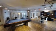 This single-floor amenity lounge is thoughtfully designed to offer a versatile social and recreational environment. The space is anchored by a full-size pool table with a rich wood frame and blue felt surface, positioned prominently at the center of the room atop warm-toned hardwood flooring that runs throughout. Track lighting mounted along the ceiling provides bright, even illumination across the entire area, complementing the clean, modern aesthetic.

To one side, a dining area features a rectangular table surrounded by contemporary white and dark chairs, suitable for casual gatherings or group seating. On the opposite end, a comfortable lounge zone offers softer seating for relaxed conversation or unwinding. A series of glass-paneled partition walls and doors define distinct areas within the open floor plan, allowing light to pass through while maintaining a sense of organization and flow.

The walls are finished in crisp white, and the overall design palette is neutral and refined, lending the space a polished, professional atmosphere. Additional details such as recessed lighting, a clearly marked exit, and well-maintained fixtures contribute to the functional yet stylish character of this communal space, making it well-suited for both entertainment and social interaction.