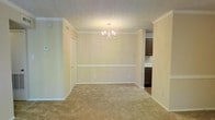 This single-floor residence offers a welcoming and functional layout with classic design details throughout. The main living and dining area is finished with warm, neutral-toned carpet that adds comfort underfoot, while crisp white crown molding and chair rail trim along the walls lend a touch of traditional elegance. A chandelier with delicate arms serves as a focal point overhead, casting warm light across the space and complementing the textured ceiling. The walls are painted in a soft, light tone that keeps the interior feeling bright and airy. A hallway door provides convenient access to additional areas of the home. Adjacent to the main living space, the kitchen is visible through an open transition, featuring dark wood cabinetry that provides a rich contrast to the lighter surroundings. The kitchen offers generous cabinet storage and connects seamlessly to the dining area, making it well-suited for everyday living and entertaining. The overall ambiance of the space is comfortable and understated, with classic architectural details that give it a timeless character. The open flow between the living, dining, and kitchen areas ensures a cohesive and practical living environment throughout this single-floor home.