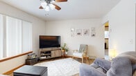 This well-organized single-floor home offers a thoughtful layout that maximizes every square foot across six distinct rooms. The living room serves as the heart of the home, featuring warm hardwood flooring, a ceiling fan, and neutral white walls that create a bright and welcoming atmosphere. A gallery wall of framed art adds a personal touch, complemented by a cozy seating arrangement centered around a large flat-screen TV on a rustic media console.

The kitchen is outfitted with white cabinetry, light tile flooring, and a full suite of appliances including a refrigerator, dishwasher, and microwave, offering practical functionality in a clean, efficient space. The bedroom provides a comfortable retreat with carpeted flooring, a window with natural light, and wall-mounted shelving for added storage. The office is a versatile secondary room with carpeted floors and a closet, suitable for work or additional storage needs.

A sun-filled sitting area with large vertical-blind windows brings in ample natural light and offers outdoor views, adding to the home's open feel. The bathroom is clean and simply appointed with a wood-vanity, framed mirror, and shower. A connecting hallway ties all rooms together, ensuring smooth flow throughout this efficiently designed home.