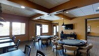 This single-floor game room is designed as a social and recreational hub, purpose-built for card games and casual entertainment. The space accommodates multiple round gaming tables, each surrounded by cushioned chairs with tan upholstery, offering comfortable seating for extended play sessions. The warm, earth-toned walls in shades of amber and brown create an inviting, lounge-like atmosphere that encourages relaxed socializing.

Overhead, a combination of pendant lights and recessed track lighting provides focused, adjustable illumination across the gaming tables, ensuring excellent visibility during gameplay. A dropped ceiling detail with intersecting beams adds architectural interest and helps define the space. Hardwood-style flooring runs throughout, contributing to the room's polished yet casual aesthetic.

Natural light filters in through several windows fitted with horizontal blinds, balancing the warm artificial lighting. A wall-mounted flat-screen television is positioned on one end of the room, alongside a dark wood console or credenza that offers additional storage and display surface. The overall layout is open and functional, making the space well-suited for community or multi-player use.