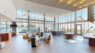 Located at 3333 S La Cienega Blvd in Los Angeles, this two-floor amenity space is defined by its striking contemporary architecture and an abundance of natural light streaming through floor-to-ceiling glass curtain walls. The main level opens into a grand, double-height lobby featuring sculptural pendant light fixtures, large-format tile flooring, and an expansive layout furnished with colorful, modern seating arrangements that encourage social interaction. Towering windows frame courtyard and pool views, connecting the interior seamlessly to the outdoors. A fully equipped kitchen anchors one wing, outfitted with warm wood cabinetry, white quartz countertops, a wine refrigerator, draft tap, and professional-grade appliances — ideal for entertaining. Adjacent dining and bar seating areas sit beneath a bold geometric gold-toned overhead installation and a vibrant patterned accent wall. The second level continues the lively atmosphere with dedicated recreation areas, including a billiards room with dual pool tables, a wall-mounted Scrabble board, and a lounge area accented by deep navy paneling decorated with colorful ping-pong paddle art. Throughout, the design balances bold accent colors with natural wood tones and ample greenery, creating a dynamic yet welcoming communal environment.