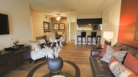 This single-floor residence showcases an open-concept layout that integrates the living, dining, and kitchen areas into one cohesive and functional space. Rich hardwood flooring flows throughout, lending warmth and visual continuity across all zones. The living area is anchored by a round glass coffee table and features a mix of seating, including a plush sofa and accent chair, complemented by tasteful wall art and decorative elements. A ceiling fan overhead provides both practical airflow and a finishing architectural touch. Adjacent to the living area, a defined dining space accommodates a table and chairs, illuminated by a ceiling light fixture. The kitchen is positioned at the rear of the open floor plan and is equipped with dark cabinetry, under-cabinet lighting, and modern appliances. A raised breakfast bar with tall dark bar stools serves as a natural divider between the kitchen and the main living space, offering additional seating and a casual dining option. The overall ambiance is contemporary and inviting, with a neutral and dark color palette that creates a cohesive, well-designed interior throughout.