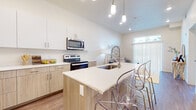 This single-floor residence showcases a contemporary open-concept design with a seamless flow between the kitchen, dining, and living areas. The kitchen is a standout feature, equipped with a full suite of stainless steel appliances including a refrigerator, range, and over-the-range microwave. Two-toned cabinetry pairs white upper cabinets with warm wood-finish lower cabinets, complemented by clean white countertops throughout. A central island with an integrated sink and seating for multiple occupants serves as both a functional workspace and a casual dining area, highlighted by stylish pendant lighting above. Soaring ceilings amplify the sense of openness throughout the main living area.

The living room benefits from abundant natural light through a large sliding glass door fitted with vertical blinds. The bedroom features soft carpet flooring and a generously sized window with blinds. A full bathroom with a wood-toned vanity and mirror is conveniently positioned adjacent to a walk-in closet with built-in shelving. Wood-look plank flooring runs throughout the main living areas, providing a warm, cohesive aesthetic. A dedicated in-unit laundry area houses a full-size Whirlpool washer and dryer, adding everyday practicality to this well-appointed space.