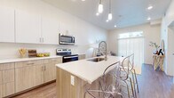 This single-floor residence showcases a contemporary open-concept design with a seamless flow between the kitchen, dining, and living areas. The kitchen is a standout feature, equipped with a full suite of stainless steel appliances including a refrigerator, range, and over-the-range microwave. Two-toned cabinetry pairs white upper cabinets with warm wood-finish lower cabinets, complemented by clean white countertops throughout. A central island with an integrated sink and seating for multiple occupants serves as both a functional workspace and a casual dining area, highlighted by stylish pendant lighting above. Soaring ceilings amplify the sense of openness throughout the main living area.

The living room benefits from abundant natural light through a large sliding glass door fitted with vertical blinds. The bedroom features soft carpet flooring and a generously sized window with blinds. A full bathroom with a wood-toned vanity and mirror is conveniently positioned adjacent to a walk-in closet with built-in shelving. Wood-look plank flooring runs throughout the main living areas, providing a warm, cohesive aesthetic. A dedicated in-unit laundry area houses a full-size Whirlpool washer and dryer, adding everyday practicality to this well-appointed space.