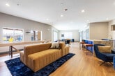 This single-floor communal lounge space is designed with a modern, open-concept layout that seamlessly connects multiple functional zones. The interior is anchored by warm hardwood-style flooring throughout, complemented by recessed lighting and clean white ceilings that enhance the sense of openness.

The main lounge area features a generous sectional sofa in rich mustard yellow, paired with deep navy blue accent chairs and plush throw pillows, all arranged atop a dark, textured area rug. A wall-mounted flat-screen TV anchors one end of the room, with a wall-mounted mini-split unit providing climate control. Adjacent to the lounge, a shuffleboard table adds a recreational element to the space.

A striking kitchenette area serves as a focal point, outfitted with bold cobalt blue cabinetry, granite countertops, stainless steel appliances including a double wall oven and microwave, and a geometric white tile backsplash. Leather-upholstered bar stools line the curved counter, creating an inviting social hub.

Further into the space, a dining and conversation area features additional seating arrangements, while a set of expansive bi-fold glass doors floods one end of the room with natural light and opens directly onto an outdoor patio. The patio, visible through the glass, features pergolas, lounge chairs, and umbrellas, extending the usable social space outdoors.