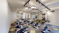 This single-floor spinning and yoga studio is thoughtfully designed to support a variety of group fitness activities in a bright, well-appointed environment. The room is outfitted with a full array of professional LifeCycle spinning bikes arranged in rows, providing ample space for participants to exercise comfortably. The flooring features a distinctive patterned carpet tile design in alternating stripes and accents, adding visual interest while offering a practical surface underfoot. Overhead, a combination of track lighting and elegant pendant light fixtures ensures the space is well-lit from every angle, creating an atmosphere that is both functional and inviting. Large mirrors line one wall, allowing participants to monitor their form and visually expand the sense of space. Floor-to-ceiling glass panel partitions and windows along the perimeter admit natural light, connecting the interior to the surrounding environment and contributing to an open, airy ambiance. The neutral wall tones and refined lighting fixtures lend the studio a polished, upscale aesthetic. Whether used for high-energy spin sessions or focused yoga practice, this versatile studio delivers a professional-grade fitness environment suited to a wide range of wellness activities.