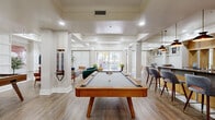 Located at 9680 W Northern Ave in Peoria, Arizona, this well-appointed single-floor community space offers a generously sized, open layout thoughtfully designed for both relaxation and socializing. The interior is characterized by warm neutral tones, durable wood-look flooring, and a coffered ceiling detail that lends architectural depth throughout.

The living area serves as a central gathering hub, featuring a wall-mounted flat-screen TV flanked by decorative panel molding, curated artwork, and an inviting seating arrangement with plush sofas and accent chairs. Large sliding glass doors and oversized windows flood the space with natural light and provide views of the outdoor surroundings.

A full-size billiards table anchors the game zone, positioned beneath recessed lighting and adjacent to a relaxed lounge nook with eclectic seating options — ideal for casual recreation and entertainment.

The kitchen area is equipped with warm wood cabinetry, dark granite countertops, stainless steel appliances, and a striking curved bar island finished with mosaic tile accents. Pendant lighting overhead adds a polished, modern touch. A dedicated beverage and snack station with a microwave and coffee equipment rounds out the kitchen's functionality.

Together, these zones create a cohesive, versatile community space that balances style with practicality.