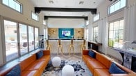 This single-floor amenity lounge is thoughtfully designed to blend modern aesthetics with comfortable communal living. The space is anchored by a striking wet bar area, accented by a vivid teal feature wall, warm stone-textured cladding, and wall sconces that cast an inviting glow. A wall-mounted flat-screen television sits centrally above the bar, making it an ideal gathering spot for entertainment. Four gold metallic bar stools line the white bar counter, adding a glamorous touch to the space.

The main lounge area features a pair of rich cognac leather sofas arranged symmetrically around a bold abstract area rug, with white sculptural ottomans serving as a stylish centerpiece. Navy blue accent pillows provide a sharp contrast against the warm leather tones. Exposed dark structural beams run across the high white ceiling, lending an industrial-chic character to the overall design.

Natural light pours in through high transom windows and large sliding glass doors on either side of the room, connecting the interior to outdoor areas and giving the space an open, expansive feel. White drapery panels frame the windows elegantly. A popcorn machine tucked near the entrance adds a fun, social dimension to the space, reinforcing its role as a vibrant hub for gathering and relaxation.