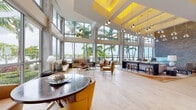 Located at 8000 West Dr in North Bay Village, Florida, this sophisticated amenity space showcases contemporary design with an unmistakable South Florida character. The grand entrance lobby greets visitors with polished terrazzo flooring, bold cylindrical pendant lighting, and a striking mosaic-tiled accent wall framing a curved reception desk — setting an elevated tone throughout.

The main lounge area is defined by soaring double-height ceilings with illuminated yellow coffers and an expansive wall of floor-to-ceiling windows that flood the interior with natural light while framing lush palm trees and shimmering waterfront views. Light hardwood flooring anchors clusters of leather seating, sleek bar-height counters, and curated artwork displayed in recessed wall niches.

A dedicated co-working and lounge area offers large format windows with sheer drapery, concrete columns, and ample long-table seating — ideal for focused work or casual gatherings. A well-appointed coffee bar features granite countertops, warm wood cabinetry, and stainless steel appliances.

Additional seating areas incorporate rich wood slat accent walls and eclectic furnishings with vibrant yellow and blue accents. Sliding glass doors open directly onto an outdoor terrace with bar seating, offering open-air views of the surrounding waterfront landscape.