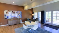 This single-floor community space serves as the welcoming hub for Azul Apartments, combining functional design with a warm, inviting ambiance. The interior is anchored by a rich, terra-cotta accent wall that adds depth and visual interest, complemented by neutral tones throughout the remaining walls and ceiling. Durable hardwood-style flooring runs across the space, providing both practicality and aesthetic continuity.

A central seating area features a round white dining table surrounded by decorative white lattice chairs, set atop a patterned area rug in muted blues and grays — ideal for casual meetings or resident gatherings. A secondary lounge area toward the back includes a pair of upholstered armchairs and a floor lamp, creating a relaxed conversation nook. Natural light streams in through large French-style doors fitted with dark curtain panels, offering both brightness and privacy control.

A low media credenza along the accent wall displays the Azul Apartments branding alongside lifestyle imagery, reinforcing the community's identity. Recessed lighting in the ceiling ensures even, well-distributed illumination throughout. The thoughtful layout balances open circulation with defined functional zones, making this a versatile and welcoming space for residents and prospective tenants alike.