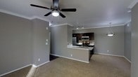 This single-floor space is designed with an open-concept layout that connects the living area and kitchen, creating a functional and cohesive environment. The living area features neutral-toned carpet flooring and is accented by crown molding that runs along the upper perimeter of the walls, adding a refined architectural detail. A ceiling fan with a built-in light fixture is centrally positioned, providing both air circulation and ambient lighting to the space.

The kitchen is well-appointed with dark espresso cabinetry that contrasts neatly against light-colored countertops. Stainless steel appliances, including a refrigerator, microwave, and range, are integrated throughout. Track lighting mounted on the ceiling illuminates the kitchen workspace effectively. A peninsula with an undermount sink extends from the main kitchen counter, serving as both a practical workspace and a natural dividing element between the kitchen and living area.

The walls throughout are finished in a consistent neutral gray tone, complementing the overall modern aesthetic. White baseboards and trim provide clean, crisp contrast. The open sightlines between rooms contribute to a spacious and connected feel throughout the entire floor plan.