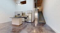 This two-story industrial loft showcases a striking blend of raw architectural character and modern finishes. The main level opens to a dramatic open-plan living and kitchen area defined by soaring ceilings that reveal exposed steel trusses, wooden ceiling decking, and industrial ductwork. Polished concrete floors reflect light throughout, enhancing the sense of volume and openness. A large ceiling fan circulates air through the expansive main living area, which features a multi-pane industrial-style window that brings in natural light.

The kitchen is thoughtfully appointed with gray shaker-style cabinetry, dark countertops, a stone tile backsplash, and a full suite of stainless steel appliances including a French door refrigerator, range, and microwave. A central island provides additional prep and gathering space. A sleek staircase with open risers connects the main level to the upper floor.

The upper level features warm wood-look plank flooring and includes bedrooms with generous walk-in closets fitted with wire shelving systems. A bathroom on this level is finished with marble-style tile surround. Hallways with polished concrete flooring connect the spaces seamlessly. Throughout, crisp white walls contrast with the industrial elements, creating a contemporary and functional living environment.