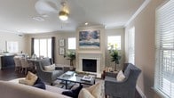 This two-story home offers a thoughtfully designed living space centered around a bright, inviting living room. The room features a vaulted ceiling with crown molding, lending a sense of height and architectural elegance. A ceiling fan with a light fixture is centrally positioned, providing both comfort and illumination. The focal point of the room is a classic white-surround fireplace, above which a bold, colorful abstract artwork adds a vibrant contrast to the room's neutral palette. Warm beige and gray tones throughout the walls and furnishings create a cohesive, calming atmosphere. Multiple windows dressed with white blinds and curtain panels allow generous natural light to fill the space, enhancing its open and airy feel. The hardwood flooring is partially covered by a patterned area rug, anchoring a seating arrangement of gray upholstered armchairs and a sofa. Potted indoor plants, including a fiddle-leaf fig, add organic warmth to the corners of the room. The overall ambiance is refined yet comfortable, blending classic architectural details with contemporary décor elements across its two floors.