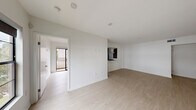 This single-floor unit showcases a clean, modern aesthetic defined by crisp white walls, light hardwood flooring, and recessed lighting throughout. The open-concept living area is bright and spacious, seamlessly connecting to a pass-through kitchen framed by a white countertop bar. The kitchen is thoughtfully designed with rich dark wood cabinetry extending to the ceiling, white quartz countertops, a stainless steel undermount sink, and a full suite of stainless steel appliances including a gas range, over-the-range microwave, dishwasher, and refrigerator. The layout provides a generous and versatile living space bathed in recessed lighting, with a second open room that offers flexible use as a living or dining area. The bedroom features a large black-framed window that frames lush greenery outside, flooding the room with natural light. A well-sized closet with modern frosted-glass sliding doors provides ample storage. Additional rooms feature the same warm hardwood floors and neutral palette, maintaining a cohesive and polished look throughout. The overall ambiance is contemporary, clean, and move-in ready, offering a functional and stylish living environment.