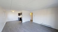 This single-floor space offers a clean, functional layout designed for comfortable everyday living. The main living area is open and airy, finished with smooth grey laminate flooring that extends throughout, creating a cohesive and modern aesthetic. Crisp white walls reflect light effectively, contributing to a bright and welcoming atmosphere. Baseboard heating units are positioned along the walls, ensuring efficient climate control throughout the space. The kitchen is partially open to the living area and features dark espresso-toned cabinetry that provides a striking contrast against the light walls. A sink and countertop workspace are visible, along with under-cabinet storage, making the kitchen both practical and visually defined. A doorway leads from the main living area into an additional room, offering separation between living and private spaces. The textured ceiling adds a subtle architectural detail overhead. Overall, the space presents a straightforward and tidy interior with a neutral color palette, functional heating infrastructure, and a layout that balances openness with defined zones for living, cooking, and private use.