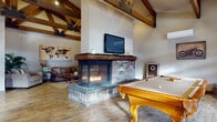 This single-floor space makes an immediate impression with its dramatic vaulted ceilings adorned with bold, exposed timber trusses that define the architectural character throughout. The open main area centers around a standout multi-sided brick fireplace with a glass surround, which serves as a focal point while seamlessly dividing the lounge and billiards areas. Rich hardwood-style flooring runs throughout, tying the various zones together cohesively.

The lounge areas are thoughtfully arranged, offering multiple seating configurations — from a casual sofa and armchair grouping near the fireplace to a more intimate reception-style sitting area featuring a navy sofa, upholstered armchairs, and a console table displaying framed artwork and informational materials. A framed gallery wall bearing the Stone Bridge branding adds a branded, professional touch.

The kitchen is well-appointed with dark cabinetry, a tile backsplash, stainless steel appliances including a full-size refrigerator, a central island with bar seating, and generous counter space. Tall windows and statement double doors with glass panels allow natural light to pour in, enhancing the warm, welcoming ambiance.

Overall, this space blends rustic charm with functional design, making it suitable for both social gatherings and professional use.