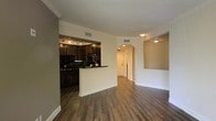This single-story space showcases an open-concept design that seamlessly connects the main living area to the kitchen and hallway beyond. Rich, dark hardwood-style flooring runs throughout, grounding the space with warmth and cohesion. Crown molding frames the ceilings, adding a refined architectural detail that elevates the overall aesthetic.

The kitchen features dark espresso-toned cabinetry paired with dark countertops, creating a bold, contrasting look. A peninsula-style breakfast bar separates the kitchen from the living area, offering both additional prep space and a casual dining option. Track lighting illuminates the kitchen workspace, while stainless steel appliances add a contemporary touch alongside the dark finishes.

A graceful arched doorway leads to a well-lit hallway, lending architectural character and guiding the eye deeper into the space. The neutral, light-toned walls throughout complement the darker flooring and cabinetry, creating a balanced and inviting atmosphere. The layout feels open and functional, with thoughtful design elements that contribute to a comfortable, modern living environment.