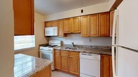 This single-floor space centers around a functional and well-designed kitchen that balances practicality with classic style. The kitchen features warm honey-toned wood cabinetry that extends to the upper walls, providing generous storage throughout. Speckled granite countertops run along the perimeter, offering ample prep space, and a matching granite island or peninsula adds additional workspace toward the front of the kitchen. White appliances, including a gas range, over-the-range microwave, and dishwasher, complement the neutral palette of the room. A stainless steel sink is neatly integrated into the countertop and positioned beneath a window, allowing natural light to filter into the space. The light-toned flooring further brightens the kitchen, contributing to an open and airy feel. The layout is efficiently arranged, with cabinetry wrapping around the walls to maximize storage and counter space, making it ideal for everyday cooking and meal preparation. Overall, the space offers a clean, timeless aesthetic with a practical design.