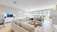 Located at 1762 Hogan Dr in Kokomo, Indiana, this single-floor space offers a thoughtfully designed open-concept layout that seamlessly integrates the kitchen and living areas. The living room is a standout feature, bathed in natural light from a sweeping wall of large windows and glass doors that frame outdoor views and create a bright, airy atmosphere. Light hardwood flooring runs throughout, complementing the warm, neutral color palette that gives the space a clean and inviting feel. A stylish fireplace with a white tile surround and wood mantel anchors one end of the living area, topped with a wall-mounted television — adding both warmth and a modern focal point. Multiple seating arrangements accommodate gatherings of various sizes, with comfortable sectional sofas and accent chairs in coordinating blue and cream tones. A dedicated dining area with a round table and upholstered chairs sits adjacent to the living space, providing a natural flow for entertaining. The kitchen is well-appointed with white shaker-style cabinetry, quartz countertops, a herringbone tile backsplash, stainless steel appliances, and ample storage. Recessed lighting throughout ensures consistent illumination, enhancing the space's clean, contemporary character.
