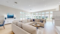 Located at 1762 Hogan Dr in Kokomo, Indiana, this single-floor space offers a thoughtfully designed open-concept layout that seamlessly integrates the kitchen and living areas. The living room is a standout feature, bathed in natural light from a sweeping wall of large windows and glass doors that frame outdoor views and create a bright, airy atmosphere. Light hardwood flooring runs throughout, complementing the warm, neutral color palette that gives the space a clean and inviting feel. A stylish fireplace with a white tile surround and wood mantel anchors one end of the living area, topped with a wall-mounted television — adding both warmth and a modern focal point. Multiple seating arrangements accommodate gatherings of various sizes, with comfortable sectional sofas and accent chairs in coordinating blue and cream tones. A dedicated dining area with a round table and upholstered chairs sits adjacent to the living space, providing a natural flow for entertaining. The kitchen is well-appointed with white shaker-style cabinetry, quartz countertops, a herringbone tile backsplash, stainless steel appliances, and ample storage. Recessed lighting throughout ensures consistent illumination, enhancing the space's clean, contemporary character.