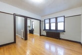 Located at 49 Dundonald St in Toronto's Church-Wellesley Village, this single-floor unit offers a thoughtfully arranged layout comprising a living room, kitchen, bathroom, and laundry area. The living room is a welcoming, open space featuring warm hardwood flooring, dark-stained wood trim and door frames, a textured ceiling, and a classic cast-iron radiator beneath a wide multi-pane window fitted with horizontal blinds. A striking white-painted brick accent wall adds architectural character, while a second radiator and additional built-in overhead cabinetry provide practical storage in a secondary nook. The compact kitchen is efficiently designed with grey laminate countertops, a stainless steel sink, white upper cabinetry, a coil-top electric range, and a window overlooking the outdoors — maximizing functionality within a modest footprint. The bathroom is a standout, featuring floor-to-ceiling large-format marble-look porcelain tile surrounding the bathtub, complemented by a natural light-filled window. The hardwood flooring runs consistently throughout the main living areas, tying the space together with warmth and cohesion. This unit combines the enduring charm of early Toronto residential architecture with practical, updated interior elements.
