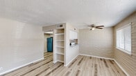 This single-floor space offers a bright, clean aesthetic defined by light wood-style flooring, textured ceilings, and distinctive painted brick accent walls that add character throughout the layout. The open living area is anchored by a ceiling fan and features a built-in shelving unit that serves as a functional room divider, creating a natural transition into the kitchen zone. A pass-through window with a granite-style countertop connects the living and dining area to the kitchen, enhancing the open feel of the space. The kitchen is well-equipped with white shaker-style cabinetry, a tile mosaic backsplash, granite-style countertops, a double-basin sink, and stainless steel appliances including a dishwasher and gas range. The bedroom provides a quiet retreat with a wide bifold closet offering generous storage and the same warm flooring found throughout. The bathroom features a tub and shower combination with tiled surround, a white vanity with a granite-style countertop, a large mirror, and a window that allows natural light to filter in. Together, the consistent finishes and thoughtful layout create a cohesive, functional living environment.