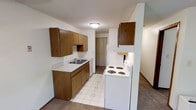 The kitchen is the focal point of this single-floor space, designed with functionality and practicality in mind. Warm medium-toned wood cabinetry lines the walls, providing generous upper and lower storage throughout. The layout features a dual-basin sink set into a light-toned countertop along one wall, complemented by a white dishwasher. On the opposite side, a white electric coil range and refrigerator complete the appliance suite, positioned conveniently near additional upper cabinetry. A partial dividing wall separates the cooking area from the adjacent space, adding subtle architectural definition to the layout. The kitchen floor is finished with a light-toned vinyl tile featuring a subtle pattern, which transitions to carpet in the surrounding areas. A flush-mount ceiling light provides overhead illumination, while a textured ceiling adds a classic touch throughout. A closet door is visible at the end of the kitchen corridor, suggesting additional storage nearby. The overall ambiance is clean, straightforward, and well-organized, with a neutral color palette that keeps the space feeling open and easy to personalize.