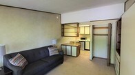 This single-floor unit offers an open-concept layout that connects the living area directly to the kitchen, creating a functional and cohesive living space. The living area features wall-to-wall carpeting and is anchored by a dark upholstered sofa accented with a patterned throw pillow. A textured, painted accent wall adds visual interest, while a wall-mounted open shelving unit with a mirrored back panel bridges the living and kitchen spaces, providing both display and storage opportunities.

The kitchen is positioned adjacent to the living area and is outfitted with light-toned cabinetry that extends to the upper walls, offering ample storage. Essential appliances include a gas range and a countertop microwave. A compact dining table with chairs sits at the boundary between the two areas, making efficient use of the available space. The kitchen features vinyl or tile flooring, contrasting with the carpeted living zone.

A closet area is visible near the entry, fitted with a hanging rod for storage. The space is illuminated by a combination of natural and artificial lighting, including a table lamp. The overall ambiance is modest and utilitarian, suited for practical, everyday living.
