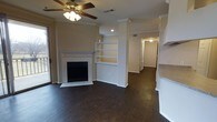 This single-floor home offers a thoughtfully designed layout with a warm, inviting ambiance throughout. The living area serves as the focal point of the space, showcasing rich dark hardwood flooring that extends seamlessly through the main areas. A classic fireplace with a tile surround and a clean white mantel anchors the room, adding both character and comfort. Adjacent to the fireplace, a set of built-in open shelving units framed by decorative columns provides functional display space while enhancing the architectural interest of the room. Crown molding runs throughout, lending a refined, polished look to the interiors. A ceiling fan with integrated lighting ensures year-round comfort and practical illumination. Natural light filters into the living area from a nearby exterior door, brightening the space. A partial view into the kitchen reveals white cabinetry and stone-look countertops, suggesting a well-appointed cooking area. A hallway leads further into the home, connecting additional rooms and maintaining a smooth, logical flow between spaces. The neutral wall tones throughout complement the darker flooring, creating a balanced and versatile interior palette suitable for a variety of décor styles.