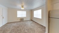 This single-floor unit offers a straightforward and functional layout designed for comfortable everyday living. The kitchen is efficiently arranged in an L-shaped configuration, featuring white cabinetry, a full suite of white appliances including a refrigerator, electric range, and dishwasher, complemented by wood-look flooring and a pass-through opening that connects to the living area. Crown molding and wainscoting paneling around the pass-through add a classic decorative touch to this space.

The living area is open and inviting, with plush carpeting underfoot, a ceiling fan overhead, and multiple windows that allow natural light to fill the room. A wall-mounted HVAC unit provides climate control. The pass-through from the kitchen creates a connected, open feel between the two spaces.

The bedroom is generously sized, also featuring soft carpeting and a ceiling fan with a light fixture. It benefits from direct access to the bathroom, which is visible through a doorway and includes standard fixtures with wood-look flooring. The textured ceilings throughout the unit lend a consistent, cohesive character to each room. Overall, the space presents a clean, functional, and well-connected single-level floor plan suited for practical, day-to-day living.