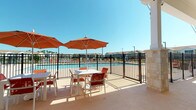 This single-level outdoor amenity space is designed to maximize comfort and leisure, centered around a large community swimming pool enclosed by a sleek black iron fence. The expansive pool deck is finished with a textured, light-toned surface and accommodates multiple dining tables paired with vibrant terracotta-cushioned chairs, each shaded by bold orange market umbrellas — ideal for enjoying sunny days outdoors.

A generous covered patio extends along the building's exterior, supported by stone-clad columns that complement the structure's mixed-material facade, which combines yellow lap siding with travertine stone accents at the base. This shaded area provides a comfortable retreat from direct sun while maintaining open views of the pool and surrounding community.

The covered lounge zone features a large sectional sofa with deep orange cushions arranged on a patterned area rug, accompanied by a wall-mounted flat-screen television — creating an inviting outdoor living room experience. Adjacent to the lounge area is a built-in outdoor grill station, adding functional convenience for outdoor entertaining. Large floor-to-ceiling windows and glass doors connect the covered patio to the interior, allowing natural light to flow freely. Palm trees and neighboring residences are visible beyond the fence, enhancing the resort-style atmosphere of the space.