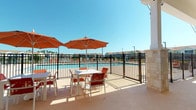 This single-level outdoor amenity space is designed to maximize comfort and leisure, centered around a large community swimming pool enclosed by a sleek black iron fence. The expansive pool deck is finished with a textured, light-toned surface and accommodates multiple dining tables paired with vibrant terracotta-cushioned chairs, each shaded by bold orange market umbrellas — ideal for enjoying sunny days outdoors.

A generous covered patio extends along the building's exterior, supported by stone-clad columns that complement the structure's mixed-material facade, which combines yellow lap siding with travertine stone accents at the base. This shaded area provides a comfortable retreat from direct sun while maintaining open views of the pool and surrounding community.

The covered lounge zone features a large sectional sofa with deep orange cushions arranged on a patterned area rug, accompanied by a wall-mounted flat-screen television — creating an inviting outdoor living room experience. Adjacent to the lounge area is a built-in outdoor grill station, adding functional convenience for outdoor entertaining. Large floor-to-ceiling windows and glass doors connect the covered patio to the interior, allowing natural light to flow freely. Palm trees and neighboring residences are visible beyond the fence, enhancing the resort-style atmosphere of the space.