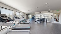 This fitness center offers a comprehensive workout environment thoughtfully arranged across a single open floor. The space is finished with durable rubber flooring throughout, providing both safety and functionality for a wide range of exercises. Large windows line multiple walls, flooding the interior with natural light and offering views of the surrounding outdoors, creating an uplifting atmosphere for training.

The cardio zone is well-appointed with multiple treadmills and elliptical trainers positioned to face the windows, allowing users to enjoy the outdoor scenery during their workouts. A dedicated strength training area features a multi-functional cable machine and a leg press/extension machine, catering to a full range of resistance exercises. A tiered dumbbell rack holds an extensive selection of free weights, complemented by kettlebells, making the space suitable for varied training styles.

A wall of mirrors enhances the sense of space and allows for form monitoring during workouts. A separate, open area is designated for stretching and floor exercises, equipped with a yoga mat mounted on the wall, a wall-mounted flat-screen television, and a small accent table. Recessed lighting and a clean, white-wall design contribute to the bright, modern aesthetic of this well-organized fitness facility.