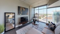 Located at 227 W Michigan Ave in the heart of downtown Kalamazoo, this contemporary urban residence blends industrial character with modern sophistication. The space is defined by its exposed concrete ceilings and structural columns, which lend an authentic loft-style aesthetic throughout.

The open living area is a standout, anchored by expansive floor-to-ceiling windows that flood the room with natural light and offer sweeping views of the Kalamazoo cityscape. The living space comfortably accommodates a seating arrangement and media setup, with the raw concrete ceiling adding texture and architectural interest overhead.

The kitchen is well-appointed with crisp white shaker cabinetry, dark granite countertops, a warm subway tile backsplash with under-cabinet lighting, and a full suite of stainless steel appliances, including a range, microwave, and refrigerator. Wood-look flooring runs throughout, tying the spaces together cohesively.

The bedroom offers a calm retreat, featuring a walk-in closet, exposed concrete ceiling consistent with the rest of the unit, and warm ambient lighting. A console table with a round mirror adds functional style to the space.

The bathroom is finished with white cabinetry, dark granite countertops, a large vanity mirror, and wood-look flooring, maintaining the refined aesthetic found throughout the residence.