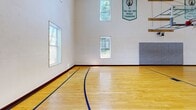 This well-appointed recreational facility in Saugus, MA offers a comprehensive range of amenities across a generously sized single floor. The centerpiece is a soaring indoor basketball court with a polished hardwood floor, regulation hoops, wall padding, and Boston Celtics championship banners adorning the bright white walls. Tall windows allow natural light to pour in, enhancing the airy, open atmosphere created by the court's impressive ceiling height. A sleek glass wall partition separates the court from the adjoining hallway, which features light hardwood flooring and a welcoming entrance with glass-panel doors.

The fitness center is a standout space, outfitted with an extensive selection of cardio machines, weight equipment, and strength-training stations. Rubber flooring in warm tones, large windows overlooking lush greenery, and ceiling fans set beneath a coffered ceiling create a bright, motivating environment. A secondary fitness area provides additional space for cycling and stretching.

The arcade room offers a vibrant retreat, featuring deep blue-gray walls, plush carpeting, foosball, pinball machines, arcade cabinets, and casual seating — ideal for relaxation and entertainment. Rounding out the facility is a functional office space, completing this versatile and thoughtfully designed recreational destination.