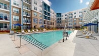 Located at 2000 Satellite Pointe in Duluth, Georgia, this outdoor amenity space is a centerpiece of a contemporary multi-story residential community. The development showcases a striking architectural design that blends warm brick exteriors with bold navy blue and white panel accents, creating a visually dynamic facade across multiple building wings. Private balconies are integrated throughout the upper floors, offering residents elevated views of the surrounding courtyard and pool area below.

At the heart of the complex lies a generously sized resort-style swimming pool with crystal-clear blue water, accessible via wide entry steps and equipped with a pool lift for accessibility. The expansive pool deck is finished in smooth concrete and lined with rows of contemporary lounge chairs and blue market umbrellas, providing ample space for sunbathing and outdoor relaxation.

A pool house structure with a striking white architectural canopy and floor-to-ceiling glass panels serves as an indoor-outdoor transition point, offering shade and shelter along the pool's edge. Additional shade structures, including a pergola-style covered seating area, are positioned throughout the courtyard. Safety amenities such as life rings and emergency signage are conveniently placed throughout the pool perimeter. Lush landscaping with manicured plantings frames the courtyard, enhancing the resort ambiance of this well-appointed outdoor retreat.