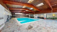 Located at 4555 Avenue Bonavista in Montréal, this remarkable single-floor indoor aquatic and leisure facility offers an impressive amount of open space designed for both recreation and relaxation. The centerpiece is a large, well-maintained indoor swimming pool with crystal-clear turquoise water, surrounded by elegant multicolored slate tile flooring that extends throughout much of the space.

The architecture is a defining feature of this facility. Soaring wood-paneled ceilings with bold, curved timber beams arch dramatically over the pool area, while multiple skylights flood the space with natural light, creating a warm and inviting atmosphere year-round. A textured natural stone wall runs along one side of the pool, complemented by built-in stone planters filled with lush greenery, adding an organic, resort-like quality to the interior.

The lounge area adjacent to the pool is furnished with wrought iron and upholstered seating, providing a comfortable space for relaxation. Classical sculptural figures are positioned poolside, lending the space an artistic and refined character. Floor-to-ceiling windows in one section of the facility frame outdoor views and allow abundant natural light to enter.

Additional utility rooms with carpeted flooring and drop-tile ceilings provide functional support space. Overall, the facility combines distinctive mid-century architectural design with practical amenities, resulting in a unique and elegant indoor aquatic environment.