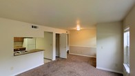 This single-floor space presents an open-concept layout that connects the living area and kitchen in a seamless, functional arrangement. The living area is finished with wall-to-wall carpeting in a neutral tone, contributing to a warm and comfortable atmosphere. A flush-mount ceiling light provides overhead illumination, while multiple electrical outlets are positioned along the walls for convenient use.

The kitchen is partially open to the living space, separated by a countertop bar that doubles as a divider and casual dining surface. White cabinetry and a full-size refrigerator are visible, maintaining a clean, cohesive look. The kitchen features hard flooring, creating a practical distinction from the carpeted living area.

A doorway visible from the living area leads to an additional room, likely a bedroom or closet space, hinting at further utility beyond the main open area. A ceiling vent ensures climate control throughout the space. The walls are finished in a soft, neutral tone with white trim and baseboards, giving the interior a simple and tidy appearance well-suited for easy personalization.