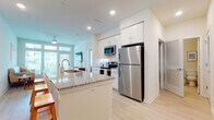 This single-floor residence is designed with a clean, contemporary aesthetic characterized by a neutral white-and-beige color palette, light wood-style flooring, and an abundance of natural light. The open-concept kitchen is a functional centerpiece, featuring white shaker cabinetry extending to the ceiling, a granite-topped island with an undermount sink and bar seating, white subway tile backsplash, and a full suite of stainless steel appliances including a refrigerator, electric range, and over-the-range microwave. A compact dining area sits just off the kitchen, accommodating a small table and chairs.

The living area is bright and welcoming, with floor-to-ceiling windows and a glass door that invite natural light and offer outdoor views of greenery. Recessed lighting and a ceiling fan add to the comfort of the space. A hallway connects the main living area to the private quarters, passing a bathroom fitted with a granite-topped vanity, white cabinetry, an undermount sink, and a large mirror. The bedroom is a serene retreat with a tall window, light flooring, and access to a walk-in closet with wire shelving. Throughout, the unit maintains a cohesive, light-filled, and efficiently designed atmosphere.