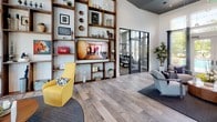 This thoughtfully designed single-floor community space offers an open, airy atmosphere highlighted by soaring double-height ceilings and an abundance of natural light streaming through large floor-to-ceiling windows dressed with dramatic dark curtains. The entry area serves as a welcoming reception and leasing office, featuring a sleek white desk workstation, a long counter workspace with bar-height seating, and a comfortable lounge corner appointed with vibrant yellow accent chairs, a round coffee table, and a textured area rug — all set atop wood-look tile flooring that runs throughout the space. A standout feature is the floor-to-ceiling custom wood shelving unit, which frames a wall-mounted flat-screen television and is styled with an eclectic mix of decorative objects, artwork, and greenery, adding warmth and personality to the room. Striking navy blue barn doors with a herringbone panel design slide open to reveal a secondary lounge or dining area beyond, offering a stylish transition between zones. Pendant globe lighting and clerestory windows above add to the bright, modern aesthetic. Adjacent to the main lounge, a glass-paneled door leads directly into a well-equipped fitness center, ensuring convenience for active residents. The overall ambiance is contemporary yet inviting, blending functional design with carefully curated décor.
