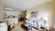 This single-floor residence offers an open-concept floor plan that integrates the living, dining, and kitchen areas into one cohesive space. The living area is anchored by a light gray upholstered sofa accented with decorative pink and patterned throw pillows, complemented by a tufted leather ottoman that doubles as a coffee table. A large abstract canvas artwork adds a focal point to the neutral beige walls, while a floor lamp provides warm ambient lighting. The carpeted flooring runs throughout the living and dining zones, contributing to a comfortable, welcoming atmosphere. The dining area accommodates a round table with seating for multiple guests, positioned adjacent to the kitchen. The kitchen features white cabinetry, a light countertop, and a breakfast bar with bar seating, separated from the dining space by a partial wall. Track lighting installed along the ceiling in the kitchen zone ensures well-lit workspaces. The overall design palette relies on soft neutrals — warm beiges, whites, and grays — creating a calm and cohesive aesthetic throughout the open living space.