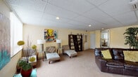 Located at Campus Edge in Carbondale, Illinois, this single-floor space serves as a welcoming common area designed for relaxed gathering and socializing. The layout is open and expansive, with soft carpet flooring running throughout and a drop-tile ceiling fitted with recessed lighting that provides consistent, even illumination across the space.

The main lounge area is furnished with a dark leather sectional sofa accented by yellow throw pillows, complemented by lounge chairs and side tables that create comfortable seating arrangements. Decorative touches include wall art, a large floor mirror with a silver frame, and accent pieces such as a teal decorative trunk and lush indoor trees that add warmth and visual interest to the pale yellow walls.

A set of elegant French doors with glass panels connects to an adjoining room, offering views into what appears to be a secondary meeting or office space. Along one wall, a pair of dark open bookshelves add functional character to the room. Near the exterior, sliding glass doors and windows with vertical blinds allow natural light to enter while providing access to an outdoor area.

Overall, the space presents a functional yet inviting atmosphere, blending practical communal design with decorative details suited to a residential community setting.