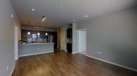This single-floor space showcases a thoughtfully designed open-concept layout that seamlessly connects the kitchen and living areas, creating a sense of spaciousness and ease of movement throughout. The kitchen is a standout feature, equipped with stainless steel appliances including a refrigerator and microwave, dark cabinetry, a subway tile backsplash, and granite countertops that extend into a peninsula-style breakfast bar. Pendant lighting and track lighting combine to illuminate the kitchen with both ambient and task-focused light. Rich, warm-toned hardwood flooring runs throughout the main living space, adding depth and character to the interior. The neutral gray walls provide a versatile backdrop that complements the dark cabinetry and modern finishes. Built-in shelving adds functional storage and display space near an interior nook. The layout includes multiple doorways leading to additional areas of the unit, suggesting a well-organized floor plan with defined zones for living and utility. Recessed ceiling vents and smoke detectors reflect a well-maintained, move-in-ready condition. Overall, the space exudes a clean, contemporary aesthetic with practical finishes suited for everyday living.