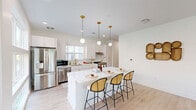 Located at 8 Duncan Ln in Arden, NC, this well-appointed single-story home offers three bedrooms, two bathrooms, and a thoughtfully designed open-concept floor plan. The heart of the home is a bright, modern kitchen featuring white shaker cabinetry, subway tile backsplash, stainless steel appliances, gold-accented fixtures, and a large center island with seating for four beneath elegant globe pendant lights. Light hardwood flooring flows seamlessly throughout, connecting the kitchen to the dining area and an airy living room bathed in natural light from oversized windows. The living room includes a ceiling fan and generous wall space ideal for art display. The dining nook offers outdoor views through large windows with stylish patterned curtains. The primary bedroom is a comfortable retreat with a walk-in closet, while additional bedrooms also feature dedicated closet space. The primary bathroom showcases a sleek white vanity with gold hardware and grab bar accessibility features. The second bathroom features a tub-shower combination with richly tiled dark walls and gold fixtures. With five closets throughout, storage is abundant. The overall aesthetic is clean, contemporary, and welcoming.