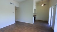 This single-floor space features an open-concept layout that seamlessly connects the living area to the kitchen. The flooring throughout is a rich, dark hardwood-style material that adds warmth and contrast to the bright, white walls. The living area is spacious and unobstructed, providing a clean and versatile canvas for furnishing arrangements.

A ceiling fan with a light fixture is centrally positioned overhead, offering both air circulation and ambient lighting to the space. The kitchen is partially separated by a half-wall partition, creating a defined yet open cooking area. White upper cabinetry lines the kitchen walls, complemented by a darker countertop surface that provides visual contrast. A refrigerator is visible at the back of the kitchen, and lower cabinetry in a dark finish adds a modern touch.

The white brick-textured wall visible in the background of the living area adds an interesting architectural detail, contributing a subtle industrial character to the otherwise clean and modern aesthetic. The overall ambiance is bright, minimal, and functional, making it well-suited for a variety of lifestyle needs.