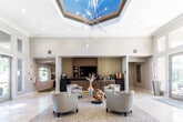 This thoughtfully designed single-story communal facility presents an impressive blend of contemporary and transitional architectural elements throughout its well-appointed spaces. The central lobby commands immediate attention with its soaring coffered ceiling accented by a striking sputnik-style chandelier suspended beneath a skylight that floods the room with natural light. The travertine tile flooring anchors the space with warmth and sophistication, while a carefully arranged seating area featuring upholstered club chairs surrounds a glass-topped coffee table, creating an inviting gathering point. Floor-to-ceiling glass doors and transom windows frame direct views of an outdoor pool area, seamlessly connecting the interior to the exterior environment.

A wide, light-filled corridor with a large arched mirror and decorative console table leads to adjacent spaces. One lounge area offers a more intimate atmosphere, furnished with a curved leather sofa, striped accent chairs, and patterned carpet, all oriented toward a dramatic floor-to-ceiling fireplace clad in large-format metallic dark tile — a striking architectural focal point. A separate dining area features a dark wood table set beneath soaring windows overlooking lush landscaping and the pool, with light hardwood flooring completing the refined aesthetic throughout.
